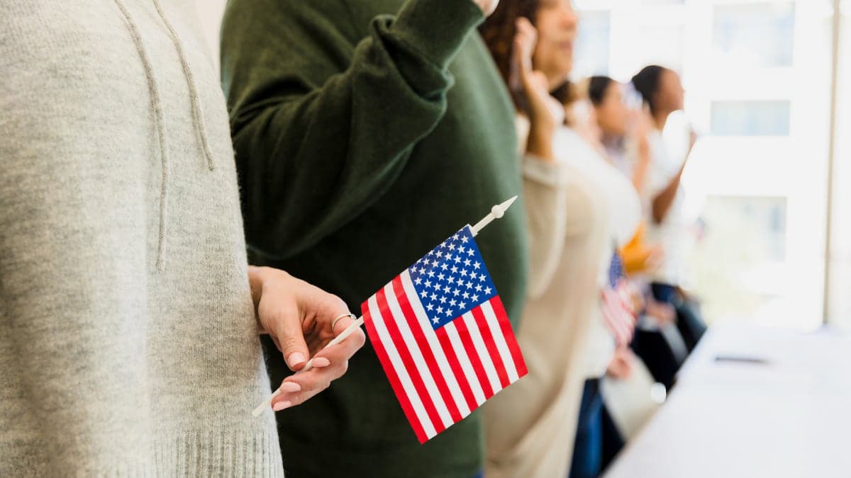 A line of people with their hands raised, taking an oath of citizenship; the first person in the foreground is holding a small U.S. flag